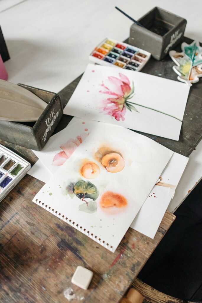 Home Artistic workspace featuring watercolor paintings and art supplies on a wooden table.